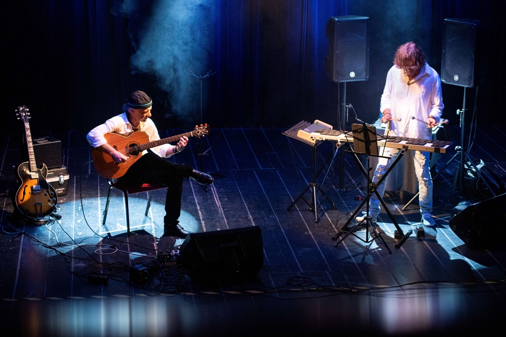 Dwóch muzyków na małej, przyciemnionej scenie grających na gitarze i wibrafonie