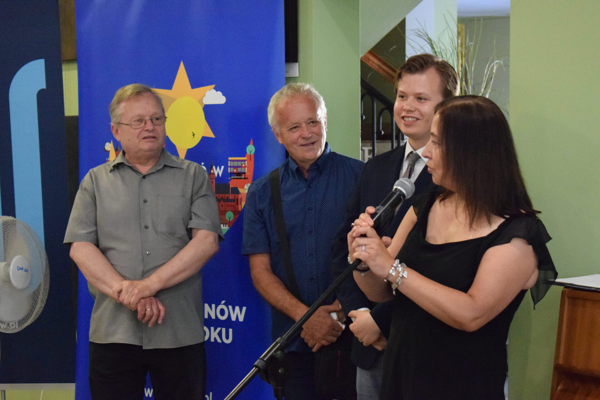 otwarcie wystawy, kobieta przemawia do mikrofonu obok stoi 3 mężczyzn