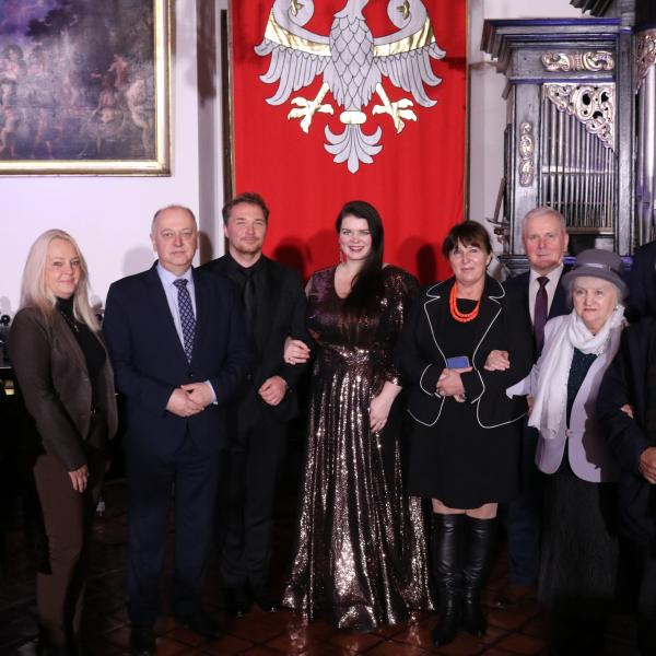 Grupa gości podczas uroczystości kulturalnej, na tle sztandaru z orłem i zabytkowych organów.