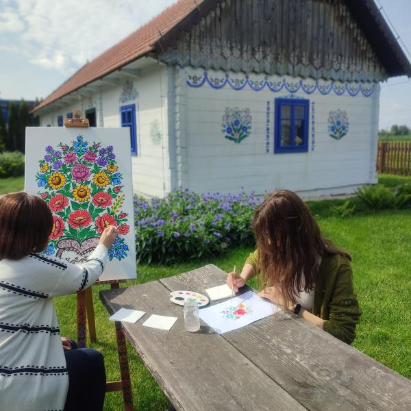 dwie kobiety malują wzory kwiatowe na tle pomalowanej białej chaty